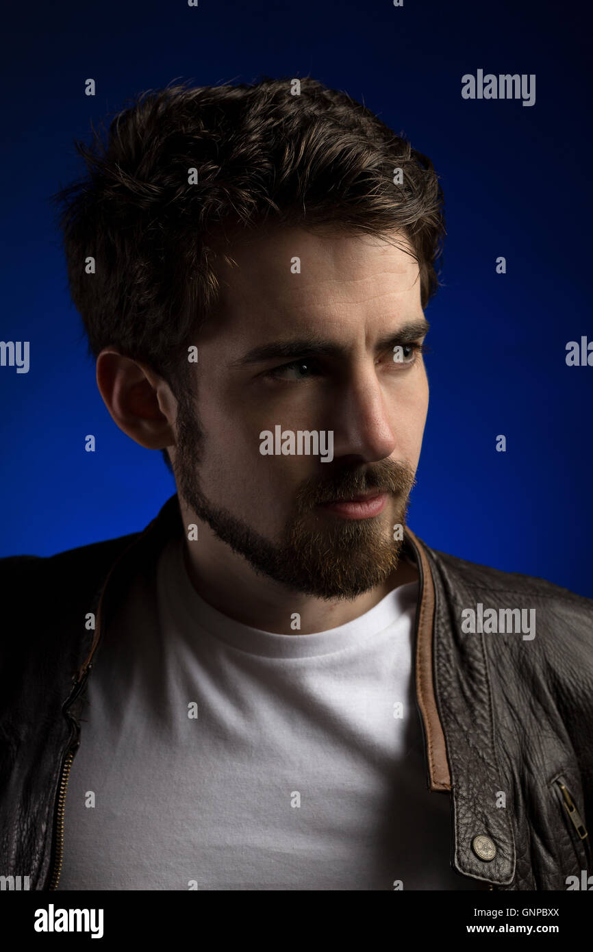 Male model with groomed beard wearing white tee and leather jacket with blue backdrop in studio Stock Photo