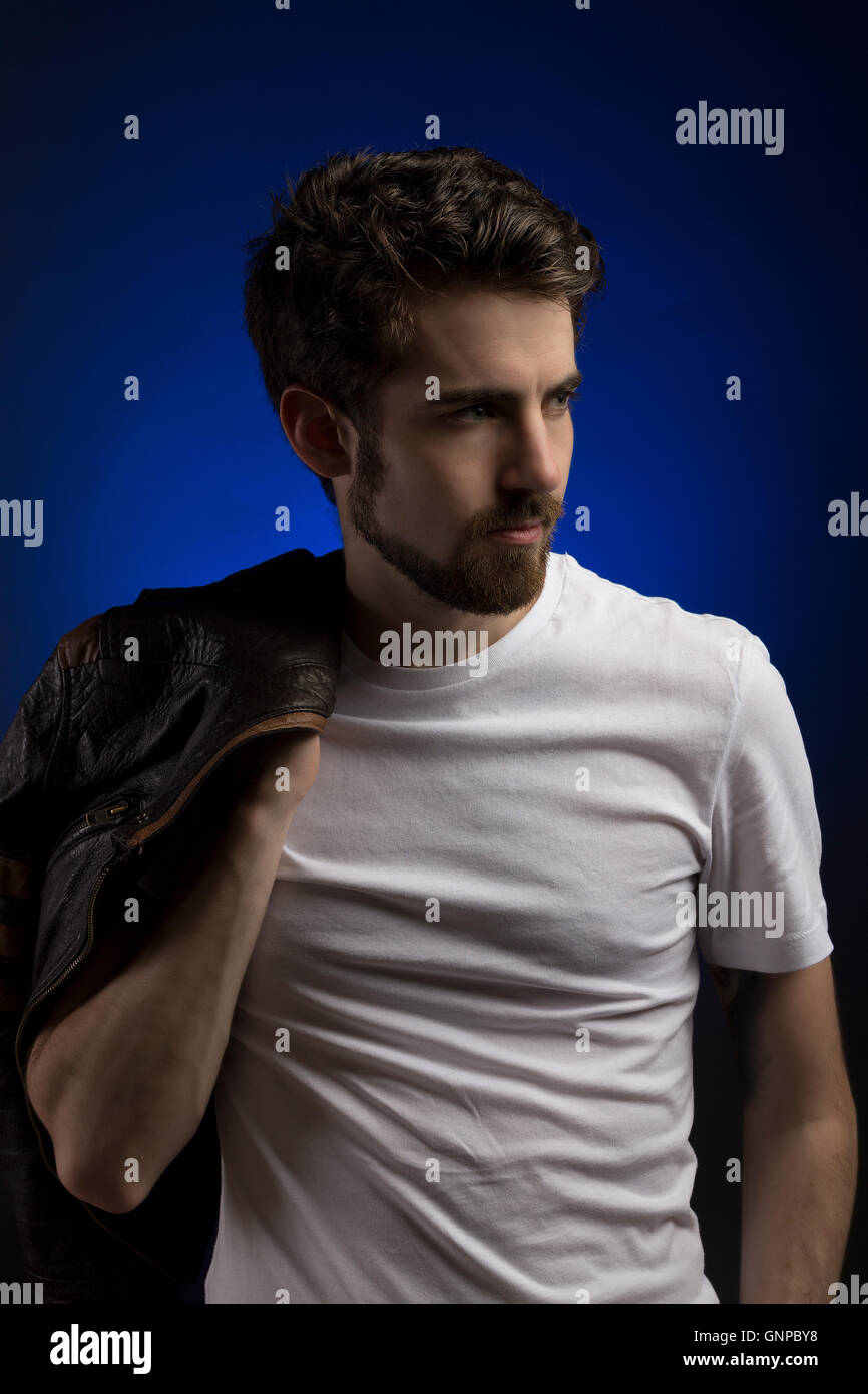 Male model with groomed beard wearing white tee with leather jacket over shoulder, blue backdrop in studio Stock Photo
