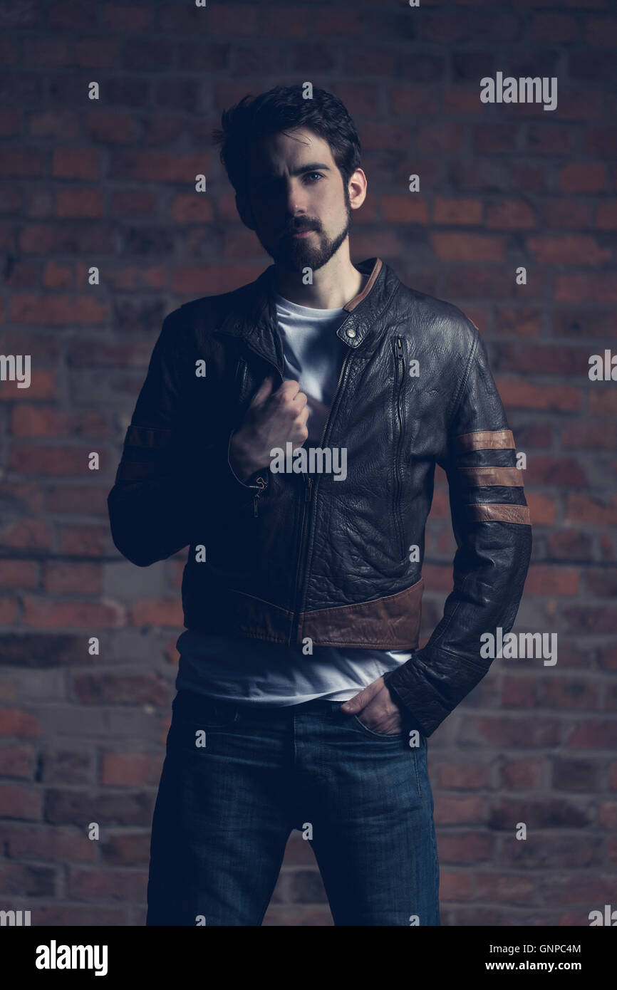 Male model with groomed beard wearing white tee and leather jacket with brick wall backdrop, shadows Stock Photo