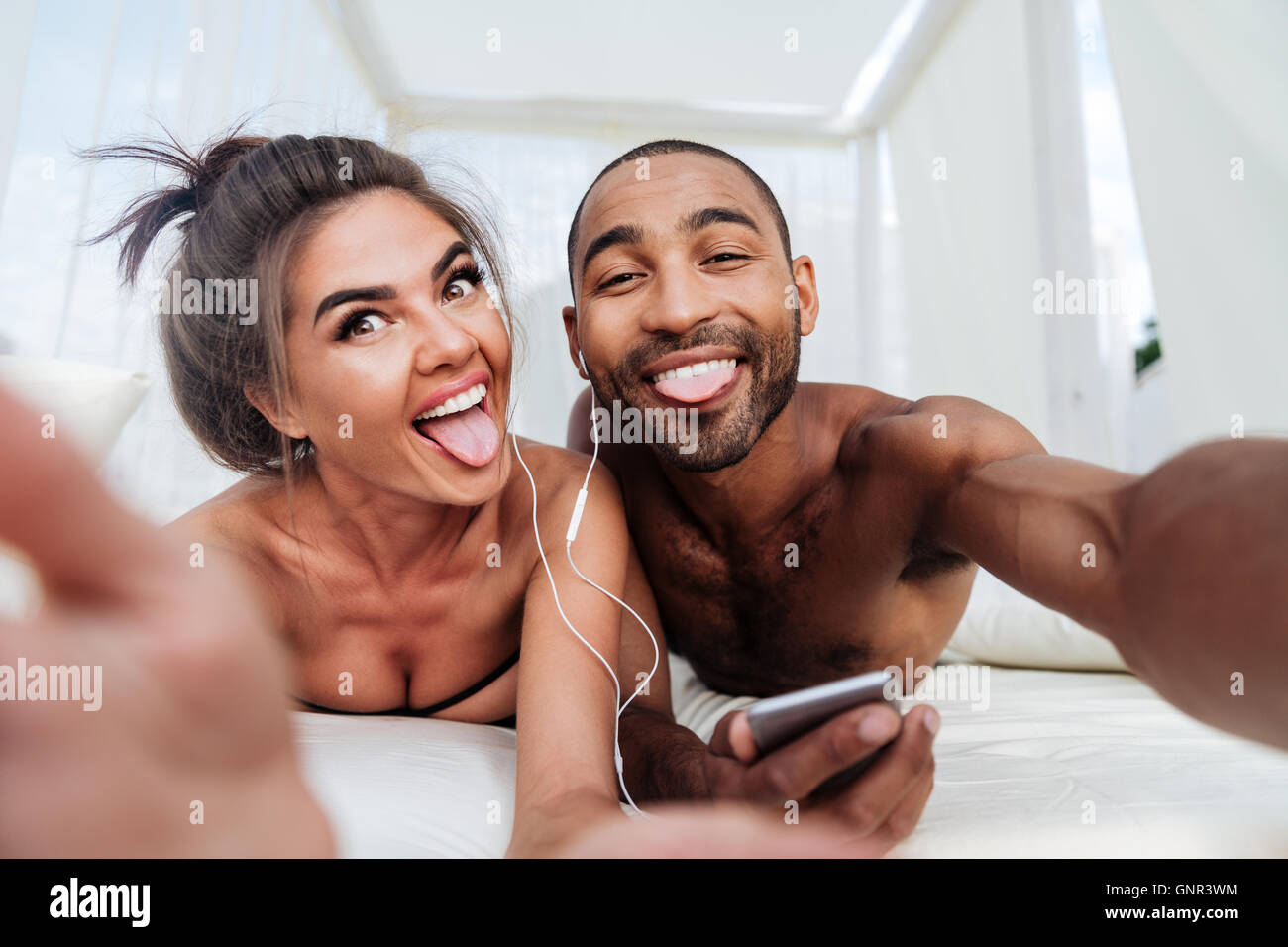 Happy cheerful couple making selfie and showing tongues while listening music at the beach Stock Photo
