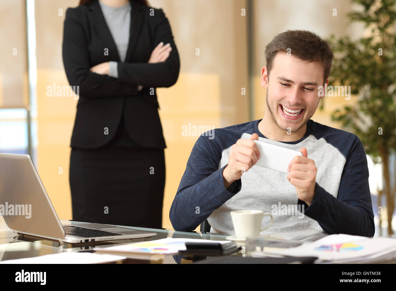 Lazy employee playing games with his smartphone sitting in a desktop while his angry boss is watching at office Stock Photo