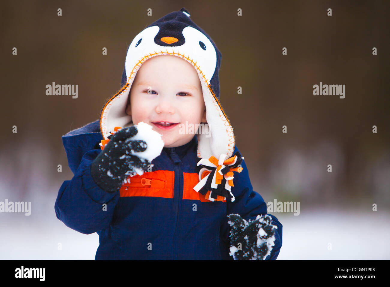 A young boy holding a snow ball and wearing a winter coat and hat smiles as he looks the camera. Stock Photo
