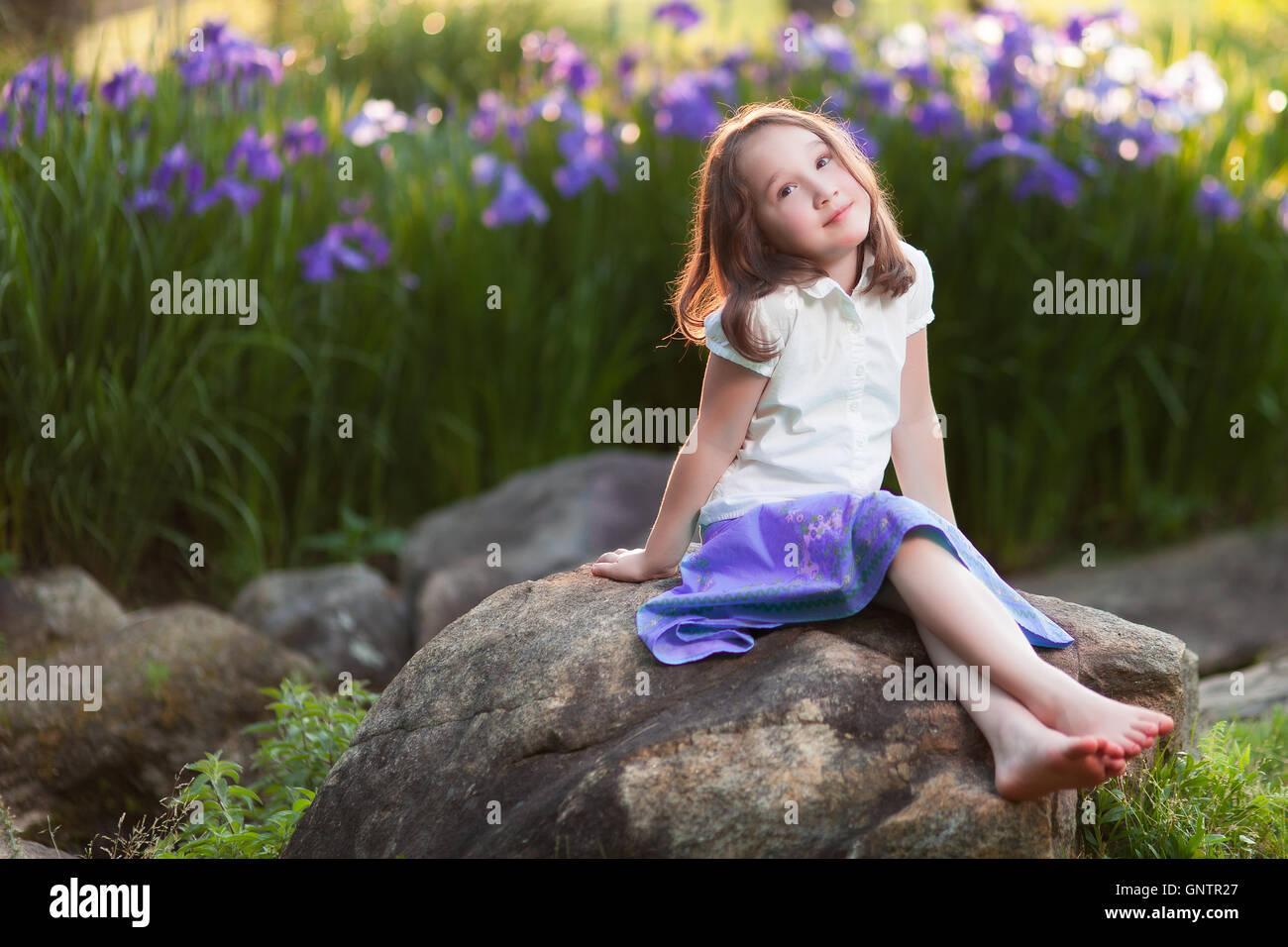 A beautiful young girl sits on a rock in a garden, deep in wistful thought. Stock Photo