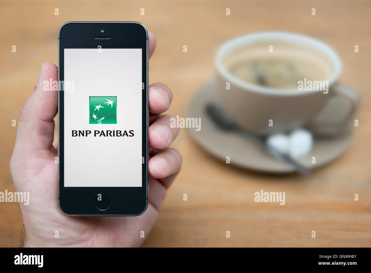 A man looks at his iPhone which displays the BNP Paribas bank logo, while sat with a cup of coffee (Editorial use only). Stock Photo