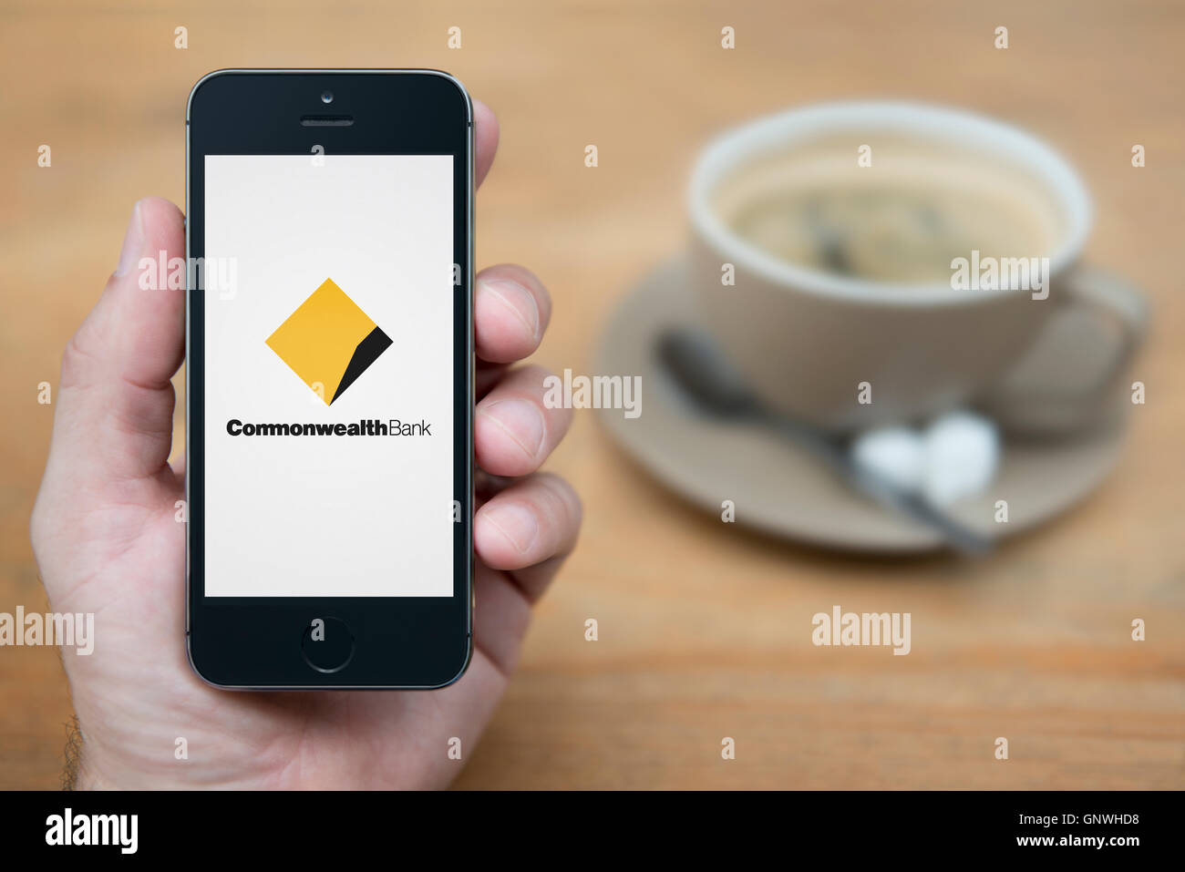 A man looks at his iPhone which displays the Commonwealth Bank logo, while sat with a cup of coffee (Editorial use only). Stock Photo