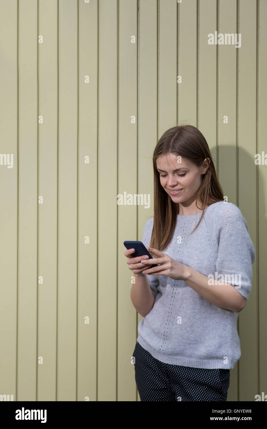 Beautiful young woman using her smart phone and leaning on a cream coloured wall. Caucasian female lifestyle concept. Stock Photo
