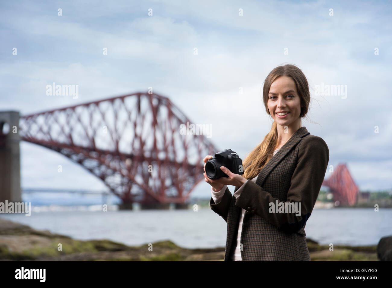 Portrait of woman taking landscape photos with a DSLR of The Forth Bridge at Queensferry, Scotland. The Forth Bridge is a UNESCO Stock Photo