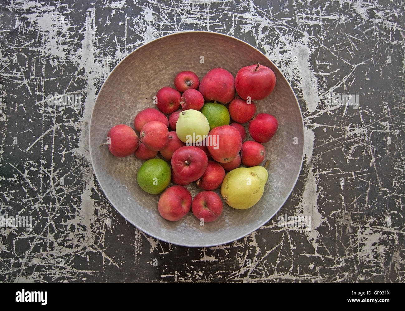 Silver color metal bowl with red apples, a pear and lime fruits on black and silver colored metal surface. Stock Photo