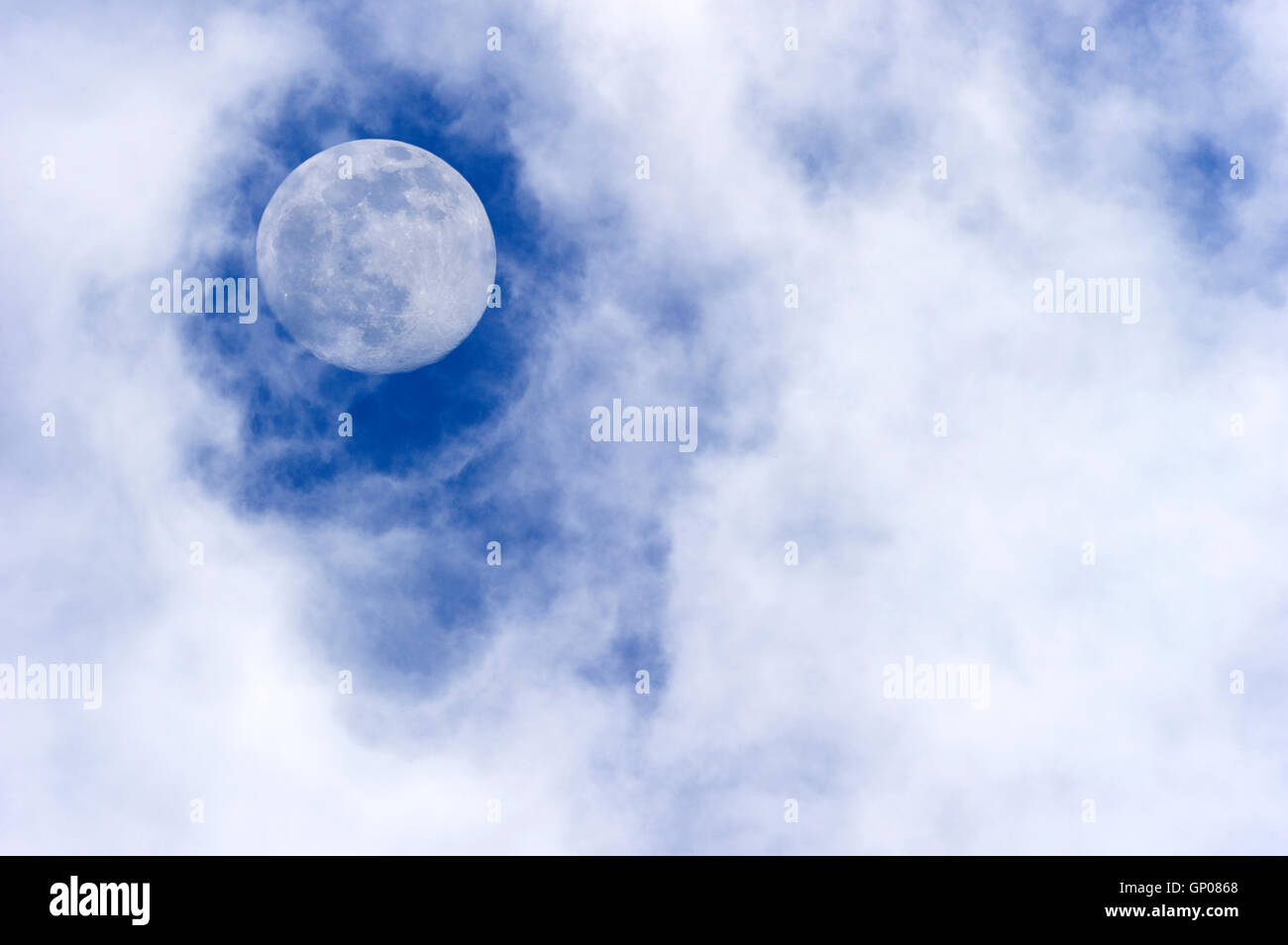 Moon clouds is a daytime scenic of wispy white cumulus clouds set against a deep blue sky as a full moon rises in the sky. Stock Photo