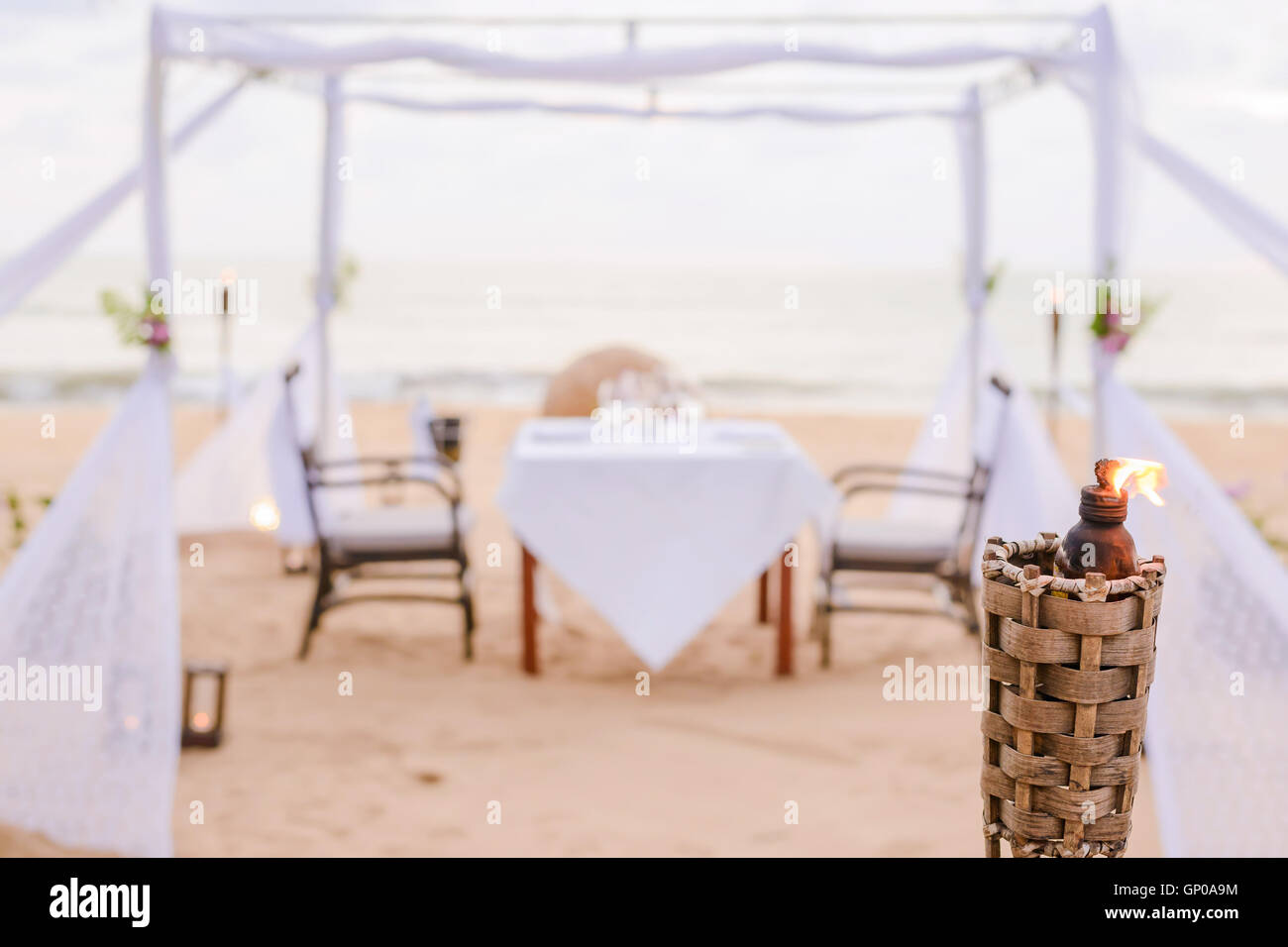 Blurry romantic dinner table setting on the beach at sunset. Focus on torch. Stock Photo