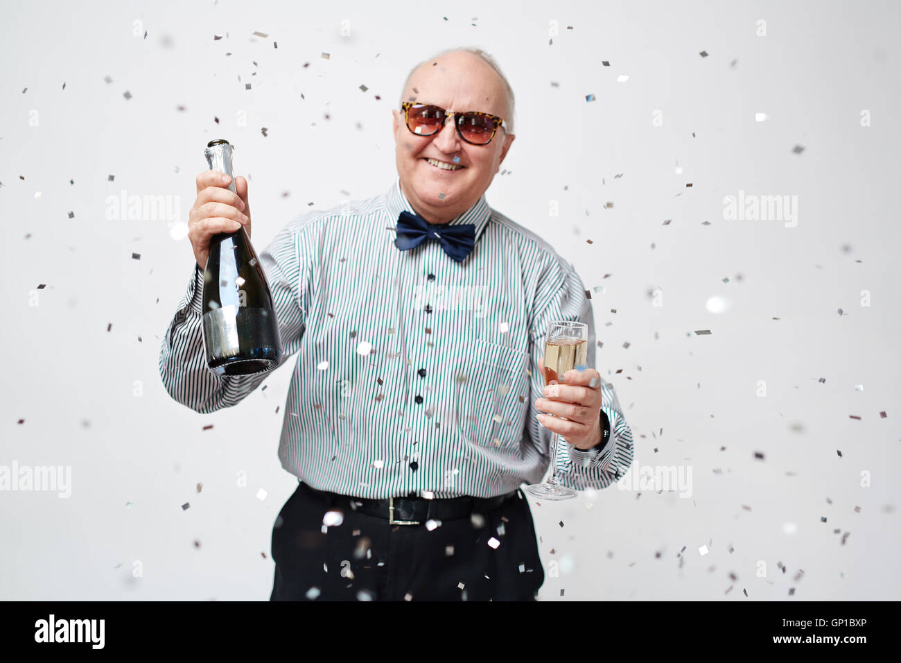 Waist up portrait of smiling well-dressed senior man in sunglasses holding bottle of sparkling wine and full wineglass with conf Stock Photo