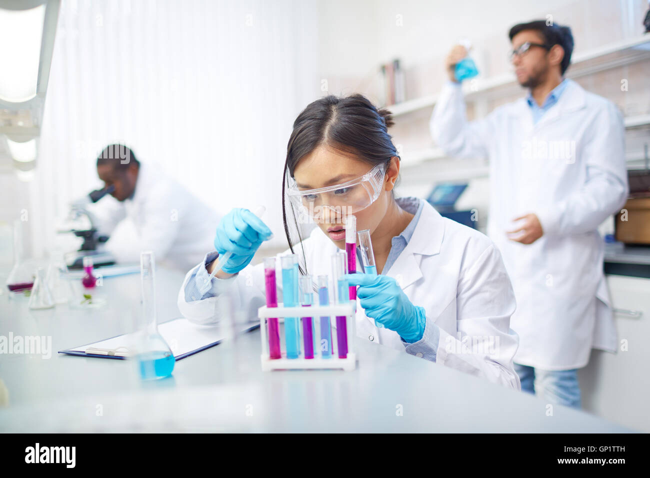 Concentrated female Asian scientist in lab coat and safety goggles working with colored test tubes in laboratory. Two colleagues Stock Photo