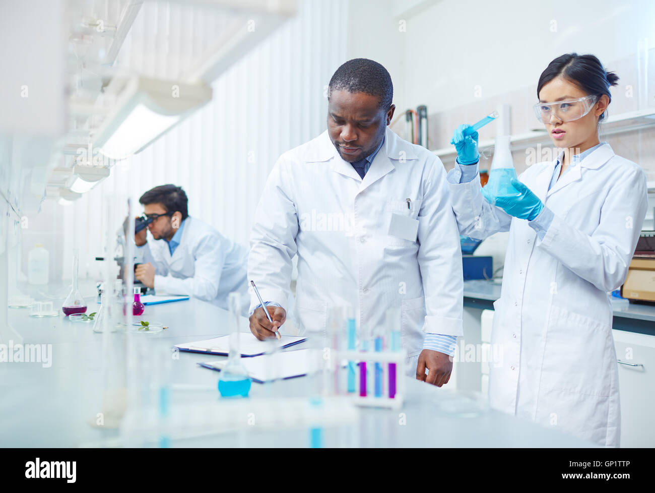 Focused female Asian scientist pouring blue liquid into flask as male African-American laboratory scientist writing down results Stock Photo