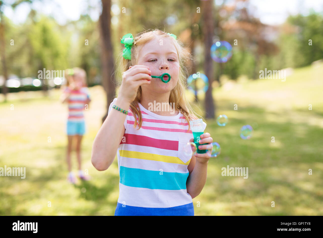 Shot of blond and freckled little girl with ponytails wearing striped t-shirt blowing bubbles in green park on sunny day. Simila Stock Photo
