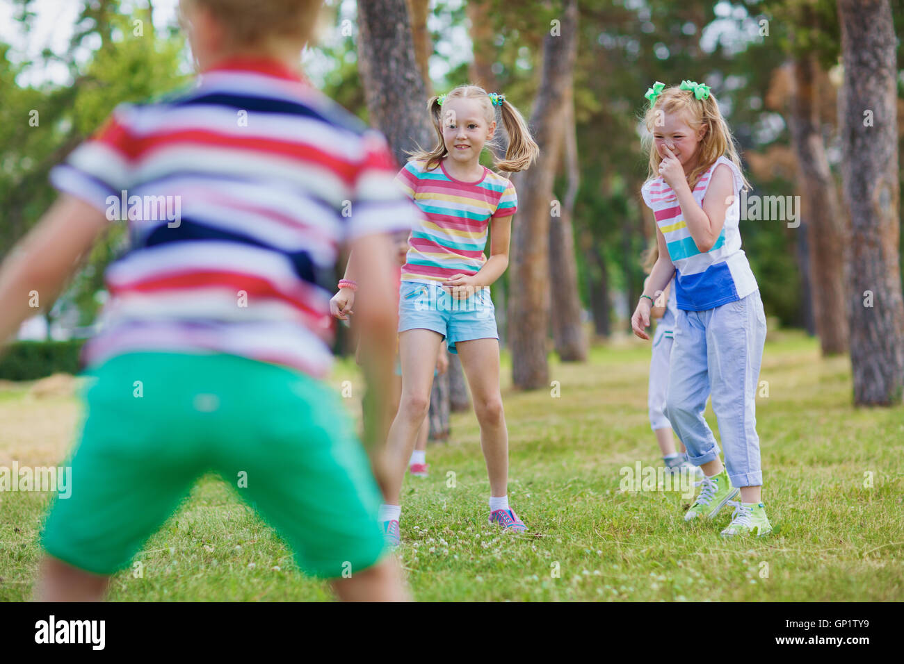 Two blond girls with ponytails in striped t-shirts playing tag in green park on sunny day with their friends. Stock Photo