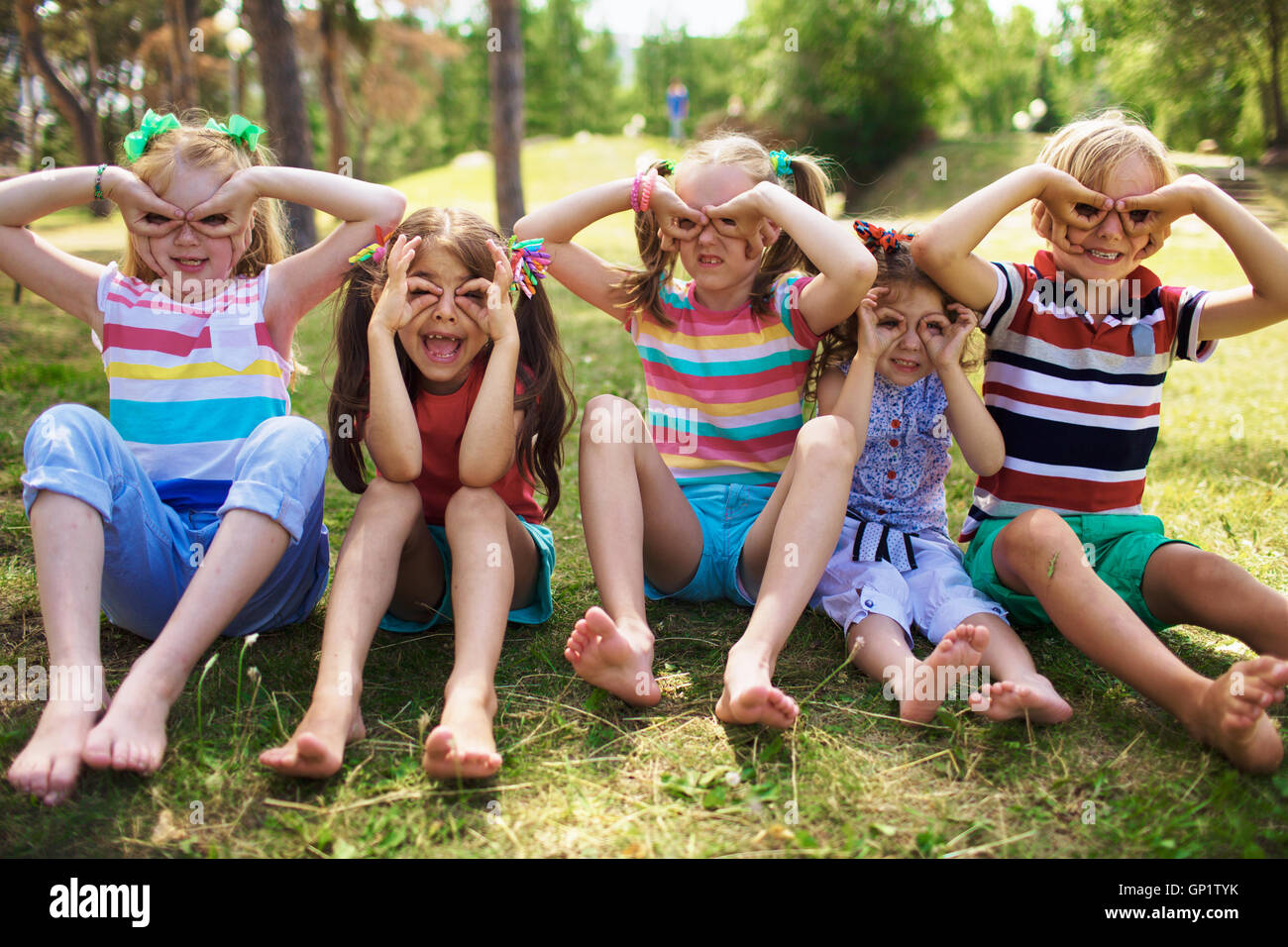 Portrait of four smiling little girls with ponytails and one little blond boy sitting on grass in green park on sunny day holdin Stock Photo