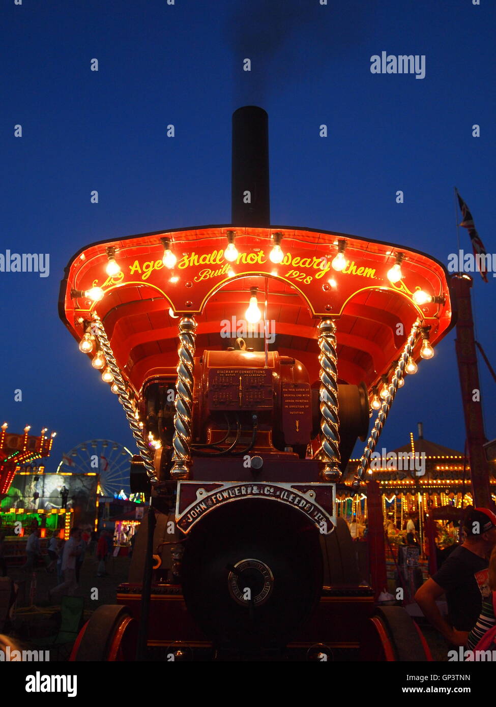 This is the front of a showmans steam traction engine lit up by its own generator and light bulbs, fun fair in the background Stock Photo