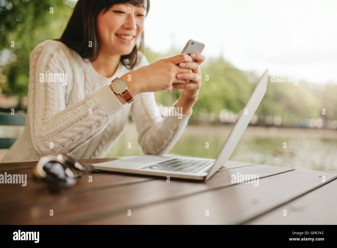Shot of smiling woman using mobile phone at outdoor cafe. Young female sitting at table with laptop reading text message on her Stock Photo