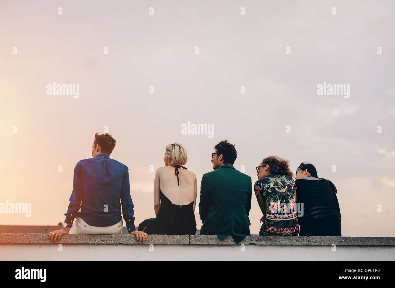 Rear view shot of young people hanging out on rooftop at sunset. Young men and women sitting on terrace in evening. Stock Photo