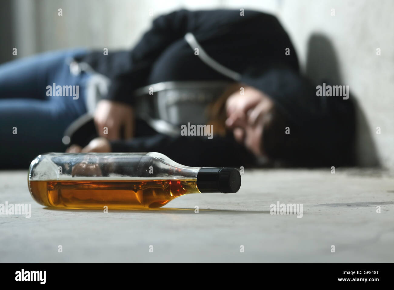 Drunk teenager on the floor in a dark and sad place and an alcohol bottle in the foreground Stock Photo