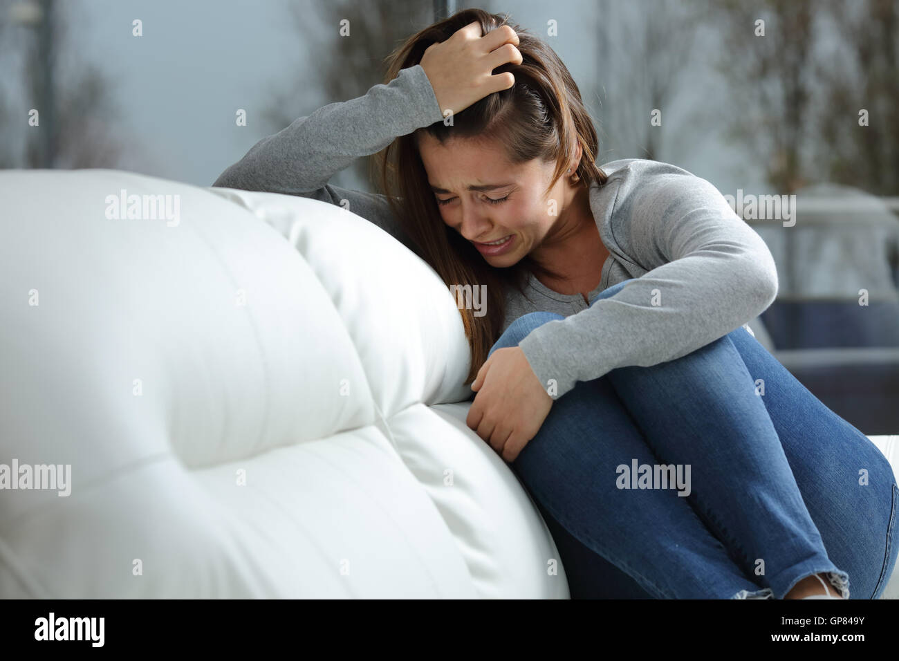 Sad girl crying desperately alone sitting on a couch at home in a dark winter day Stock Photo