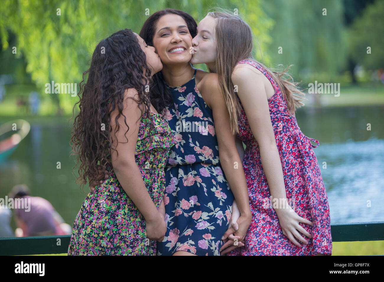 Happy Hispanic mother being kissed by two teen daughters in park Stock Photo