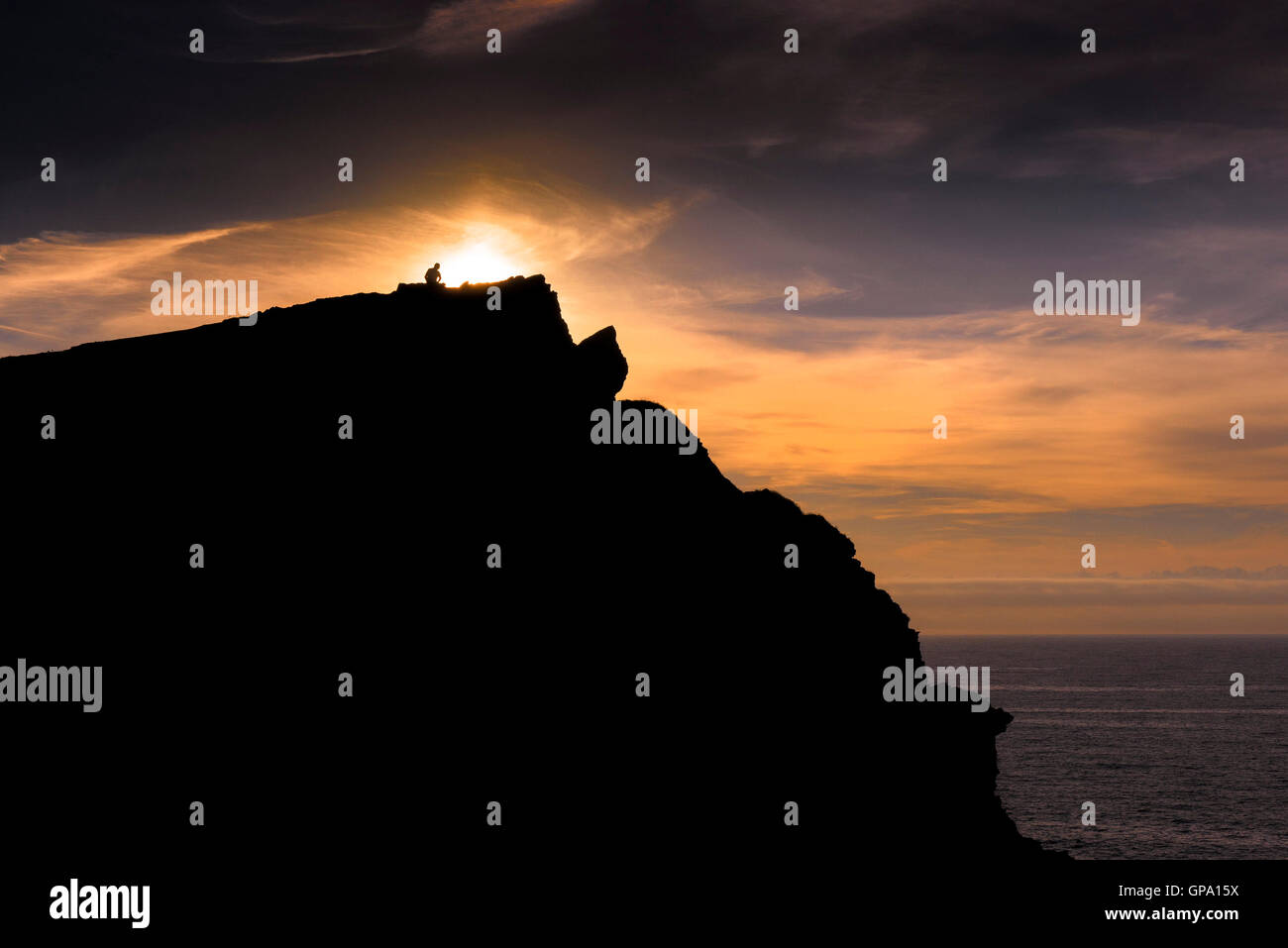 A man is illuminated by an intense sunset as he sits on the summit of Porth Island in Newquay, Cornwall. Stock Photo