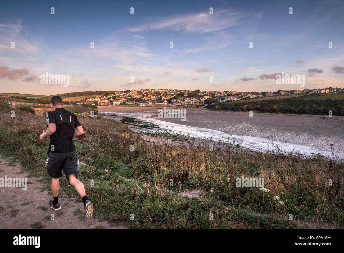 A man runs along a footpath past Porth Beach with Porth in the background.  Newquay, Cornwall. Stock Photo