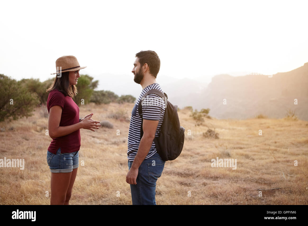 Nice young couple talking on a plato on a mountain in bright sunshine wearing t-shirts and jeans and a straw hat and backpack Stock Photo