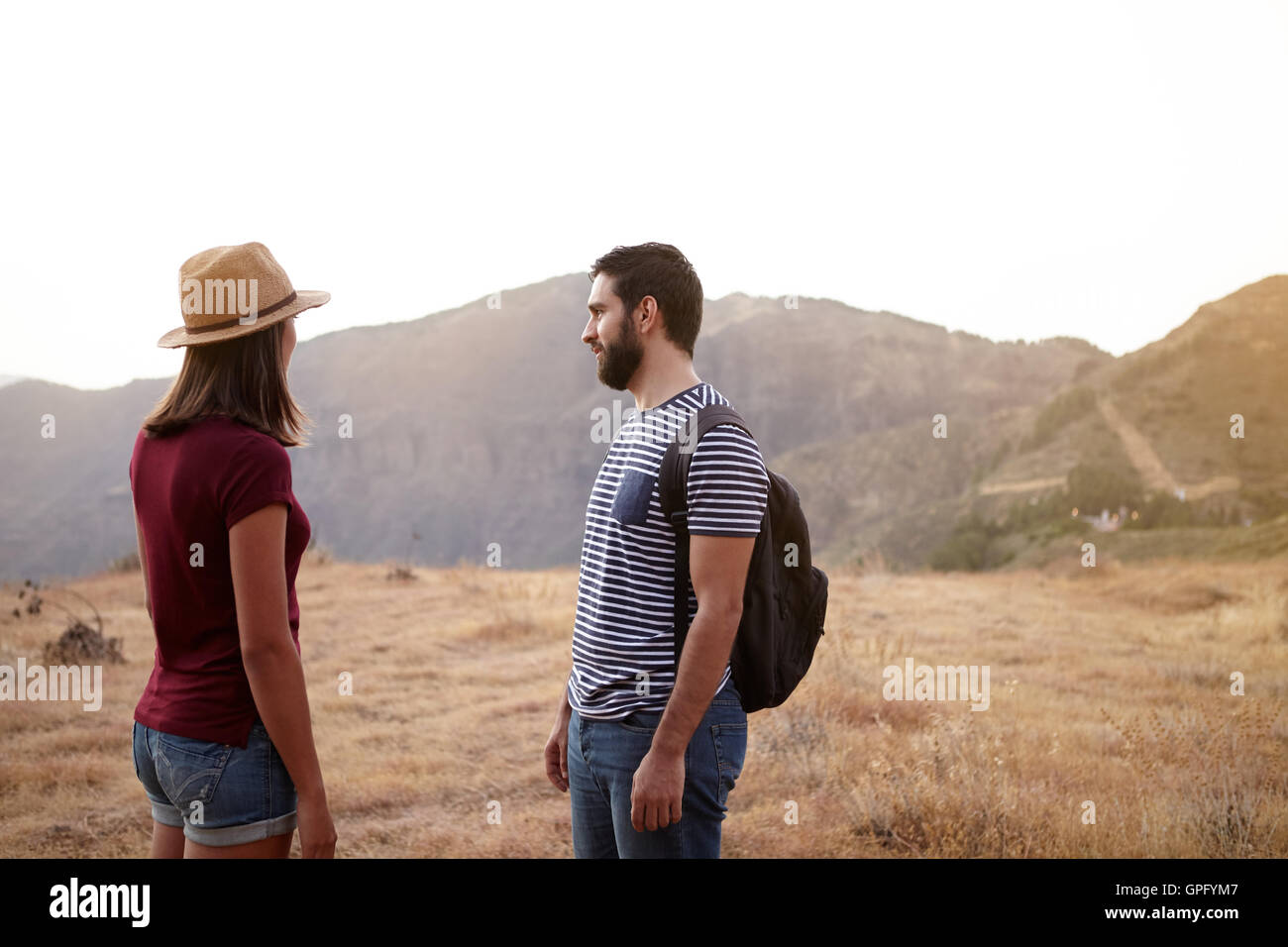 Nice young couple standing on a plato on a mountain top in bright sunshine wearing t-shirts and jeans and a straw hat and backpa Stock Photo