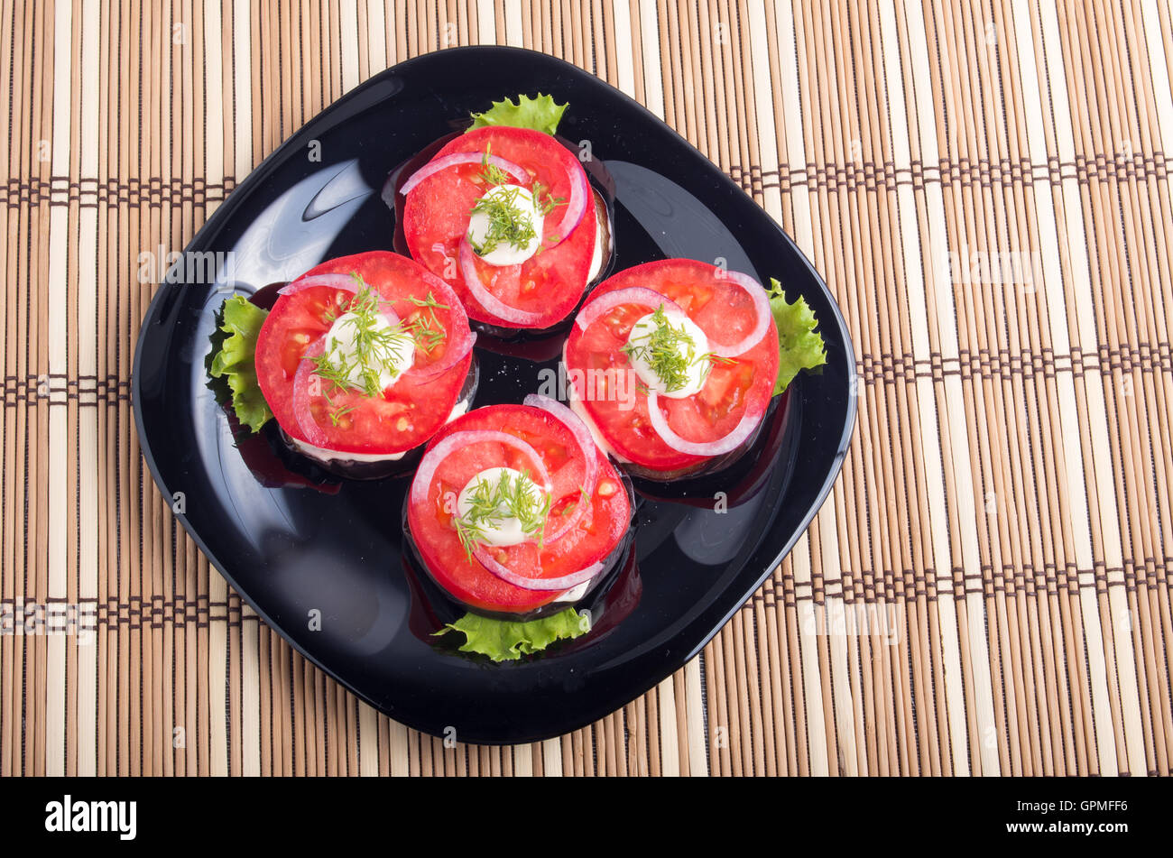 Top view of a sliced red tomatoes slices, decorated with onion and dill on a black plate on a mat made of bamboo Stock Photo