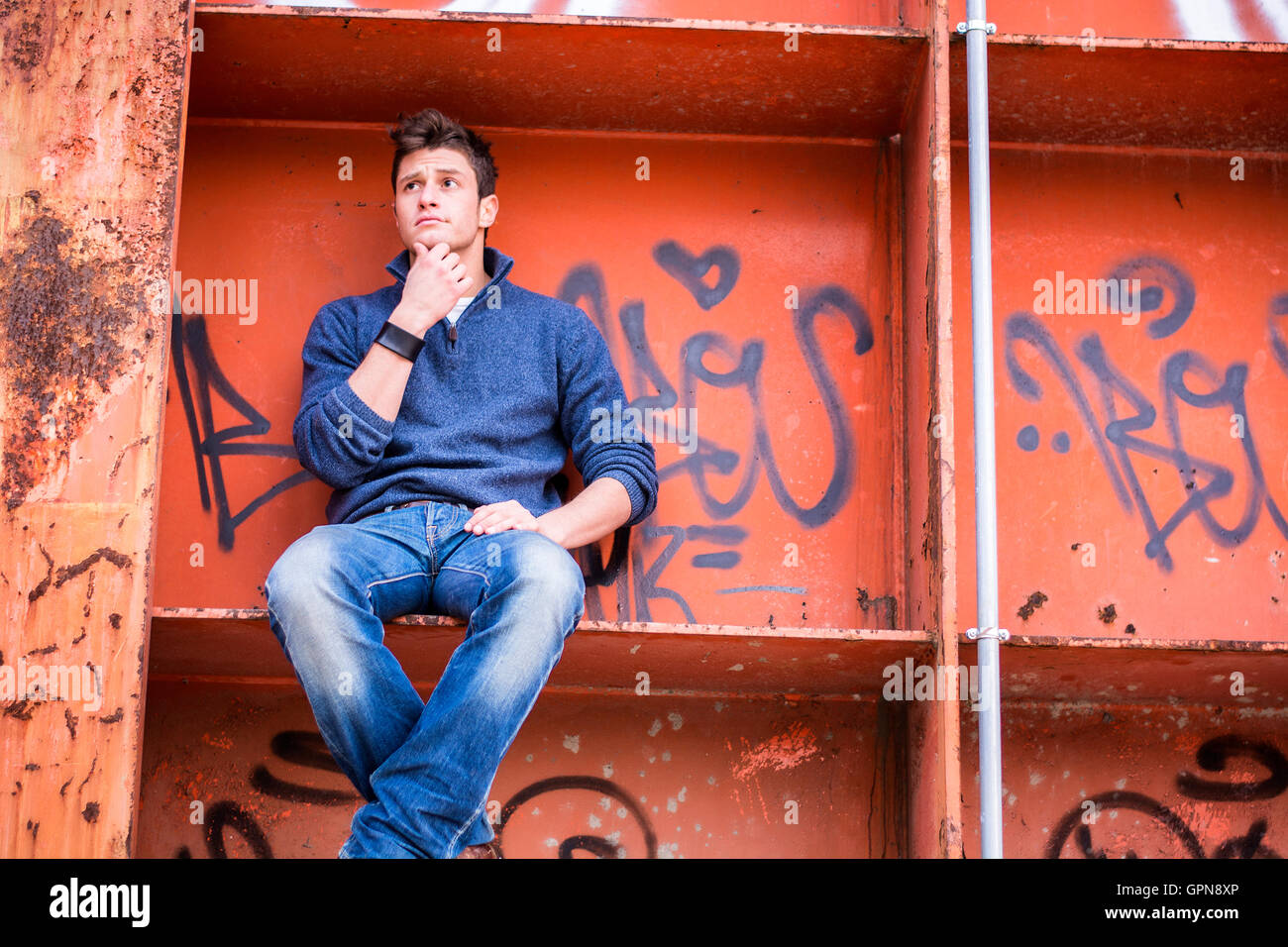 Attractive young man in urban setting sitting on orange metal structure, looking puzzled, thinking Stock Photo