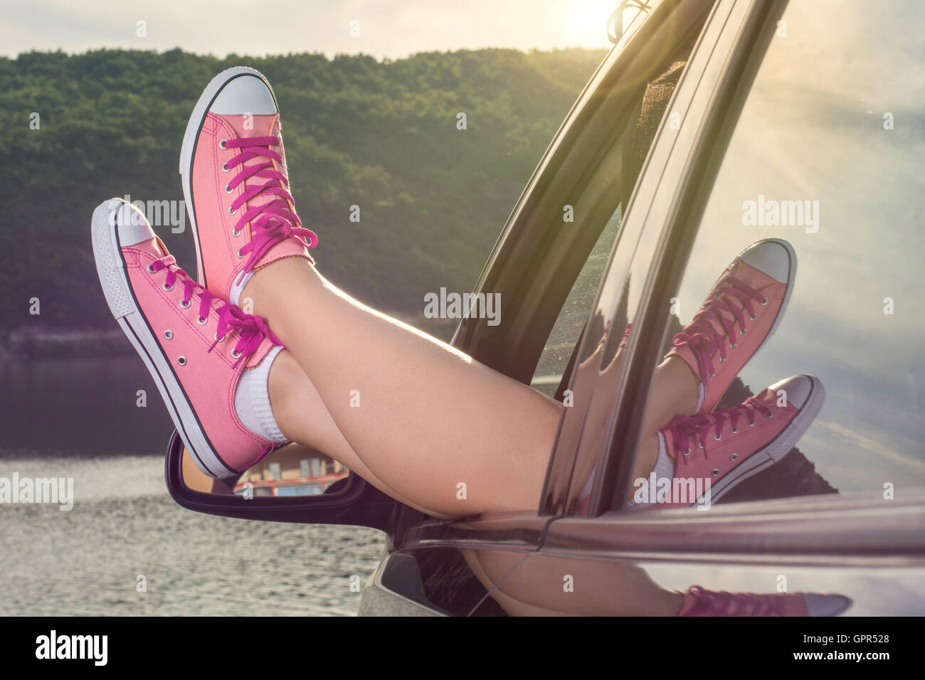 Woman legs out of the car window by the lake at sunset Stock Photo