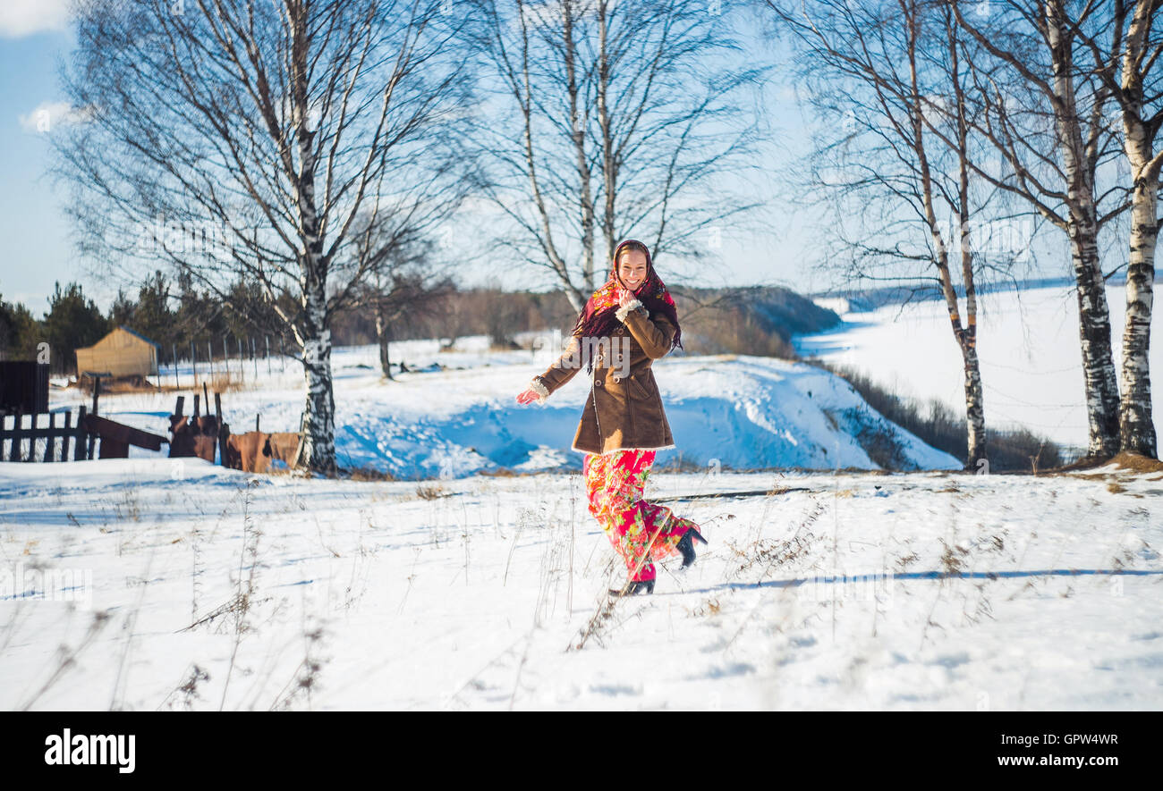 smiling young beautiful blond woman in russian winter suit runs through the snowdrifts. Girl clothed scarf, sheepskin coat and bright long red skirt Stock Photo
