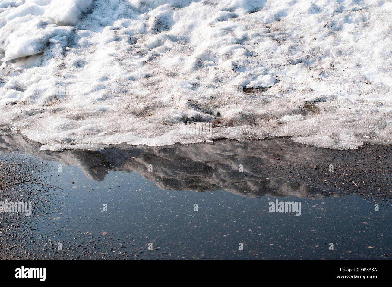 big snow heap melting on asphalt pavement with reflection in puddle