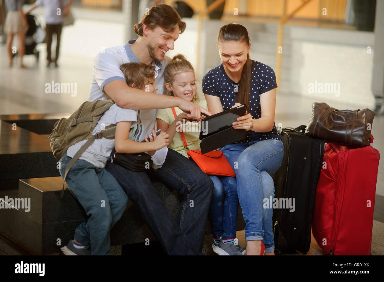 Parents with two children at the railway station. Family in large waiting room. All have settled down on bench. Mom shows someth Stock Photo
