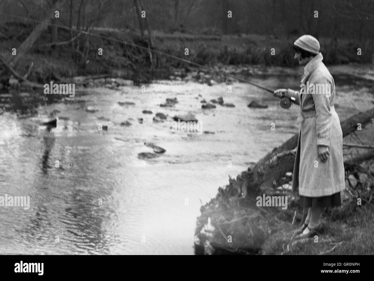 1930s, historical, an elegant young lady wearing an overcoat and hat standing fishing beside the River Rye, North Yorkshire, England. Stock Photo