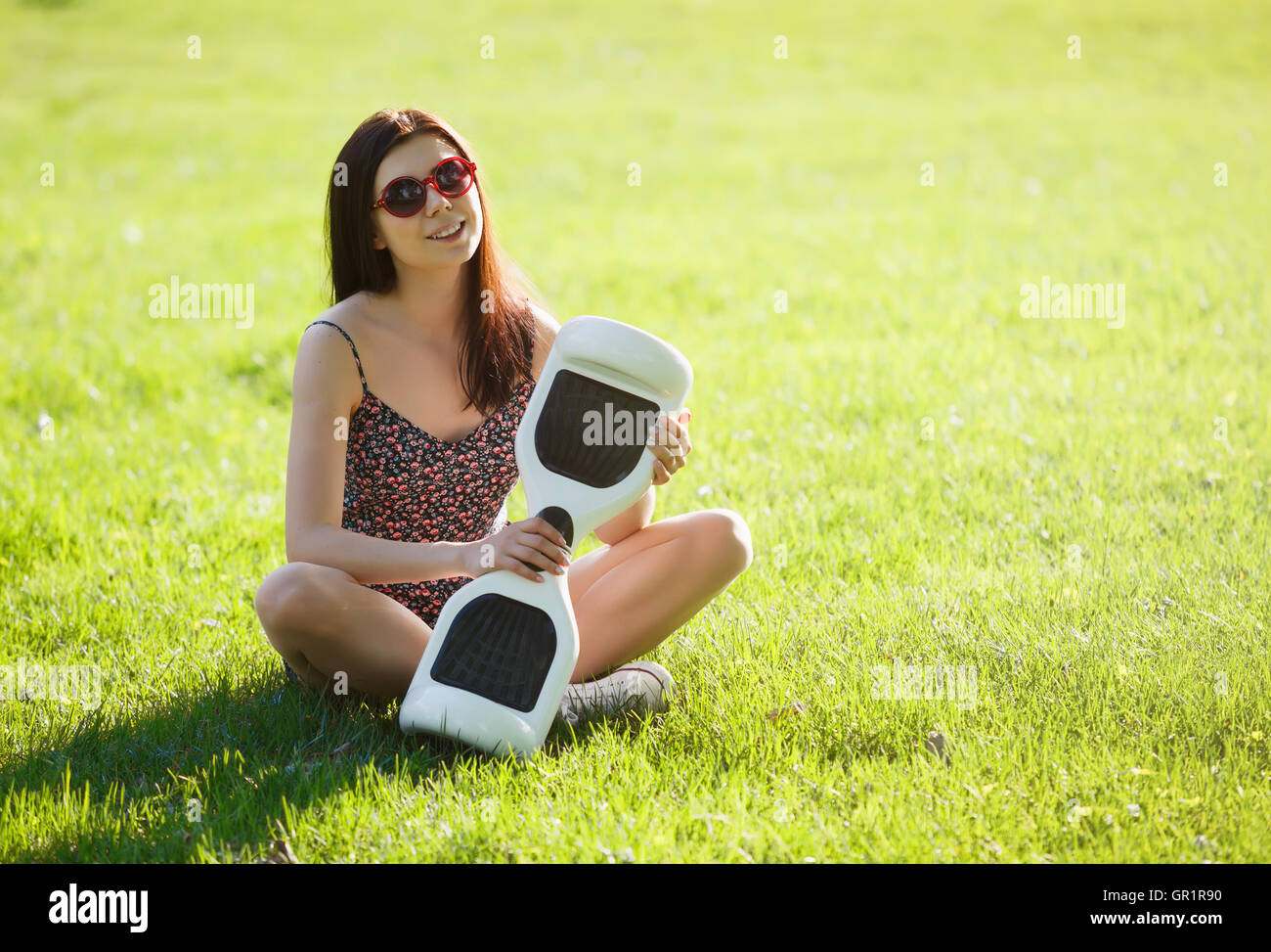 Young brunette woman with electrical mini hover board scooter in green park. Good summer weather, trendy transportation technology and cute model. Stock Photo