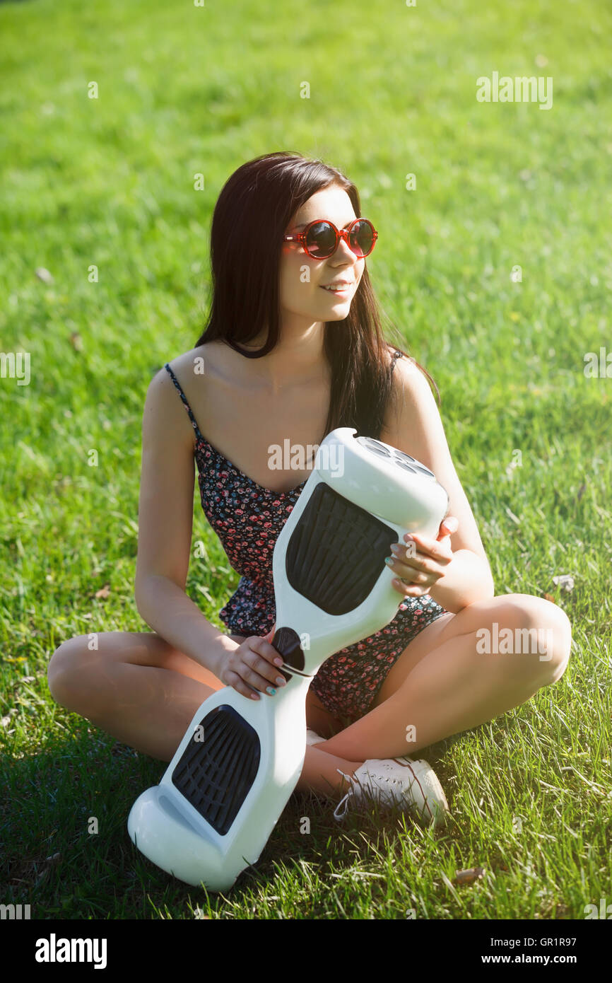 Young brunette girl with electrical mini hover board scooter in green park. Good summer weather, trendy transportation technology and cute model. Stock Photo