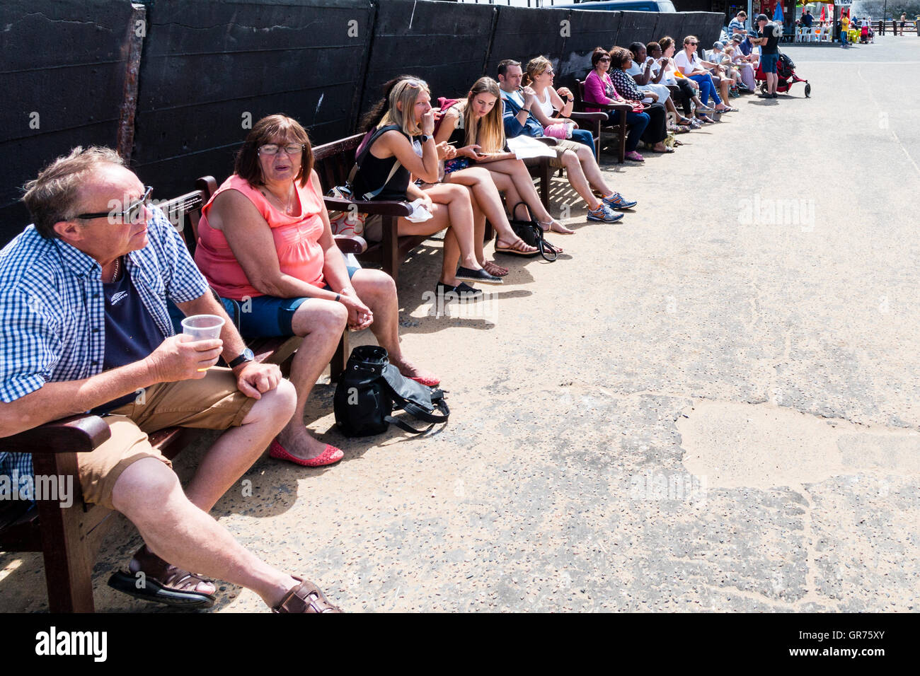 England, Broadstairs. Row of people, young and old, sitting on benches along harbour wall taking shade from the bright sunshine on a hot summer's day. Stock Photo