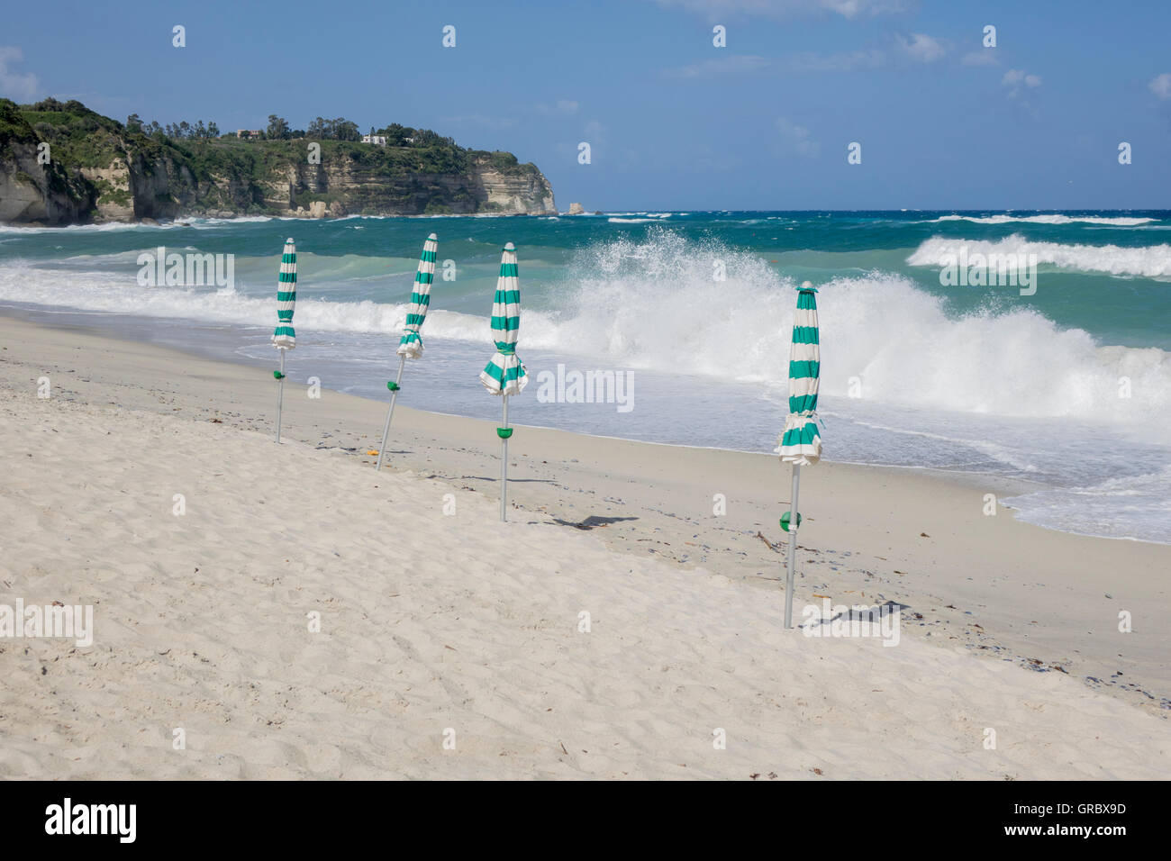 White Sandy Beach, Blue Sky, Waves, Closed Green And White Sun Shades, Cliffs With Some Trees In The Background. Tropea, Calabria, Italy Stock Photo