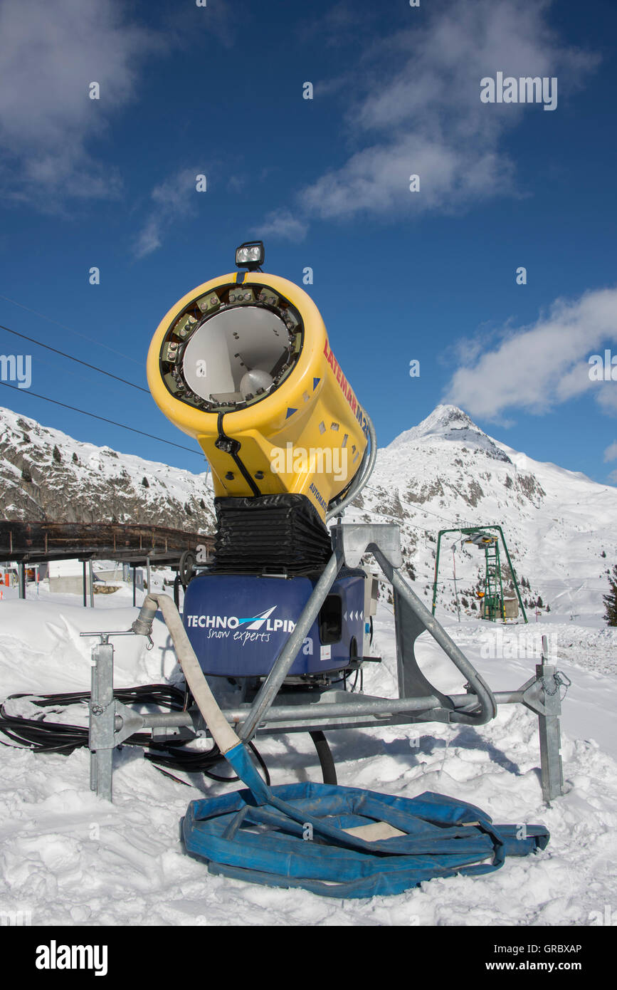 Inactive Yellow Snow Cannon Against Blue Sky, In The Background Snow Covered Mountains Stock Photo