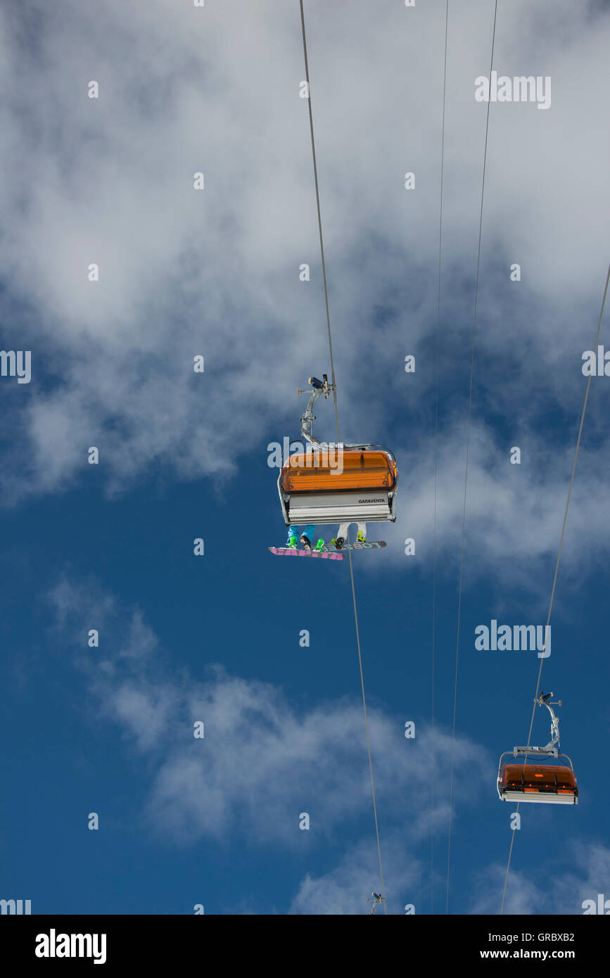 Chair Lift With Orange Weather Protectors And Two Snowoarders Showing The Lower Side Of Their Boards, Against The Blue Sky And White Clouds Stock Photo