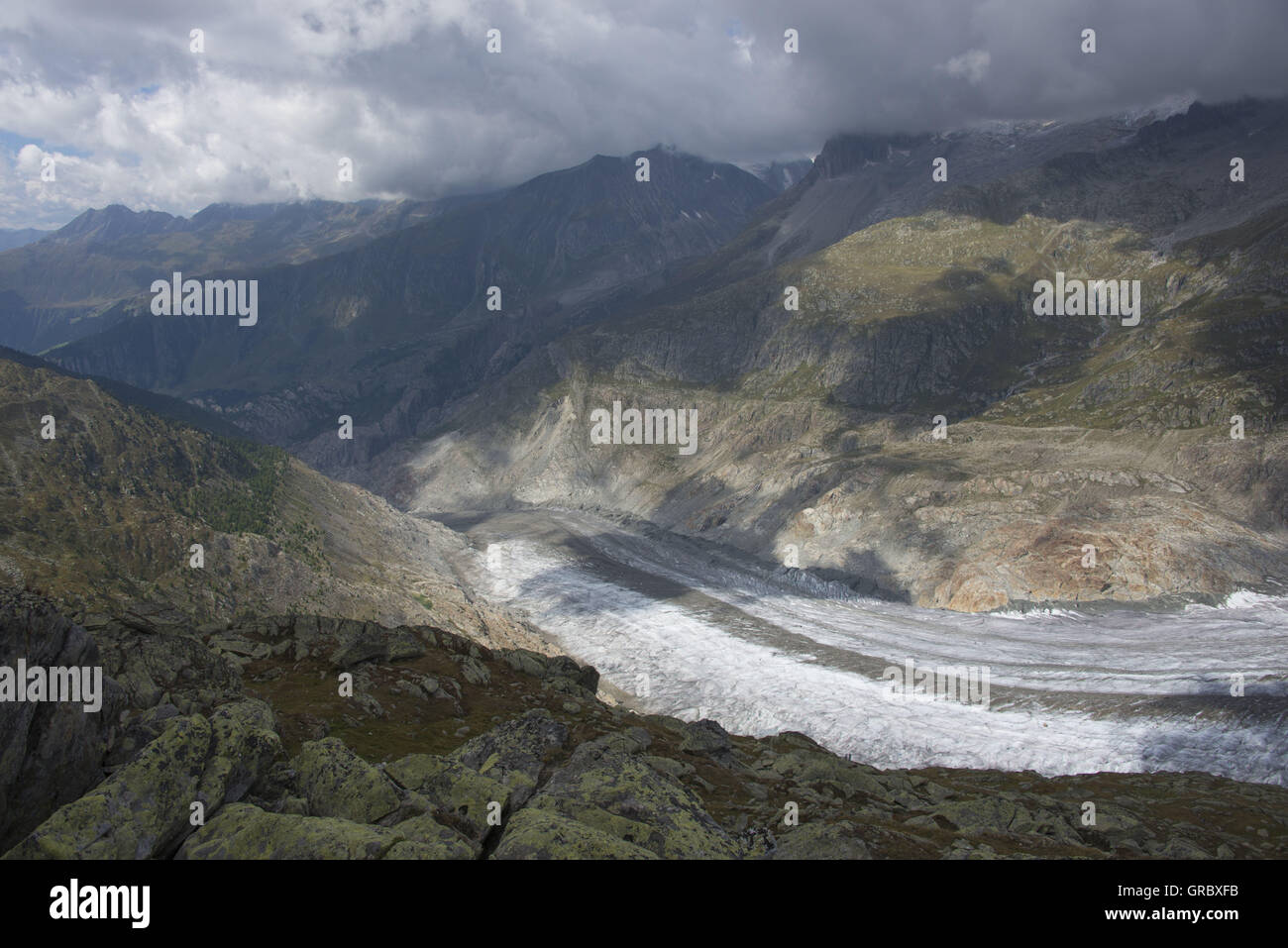 View Over Rocks On The Great Aletsch Glacier, In The Background Mountains And Dramatic Clouds Stock Photo
