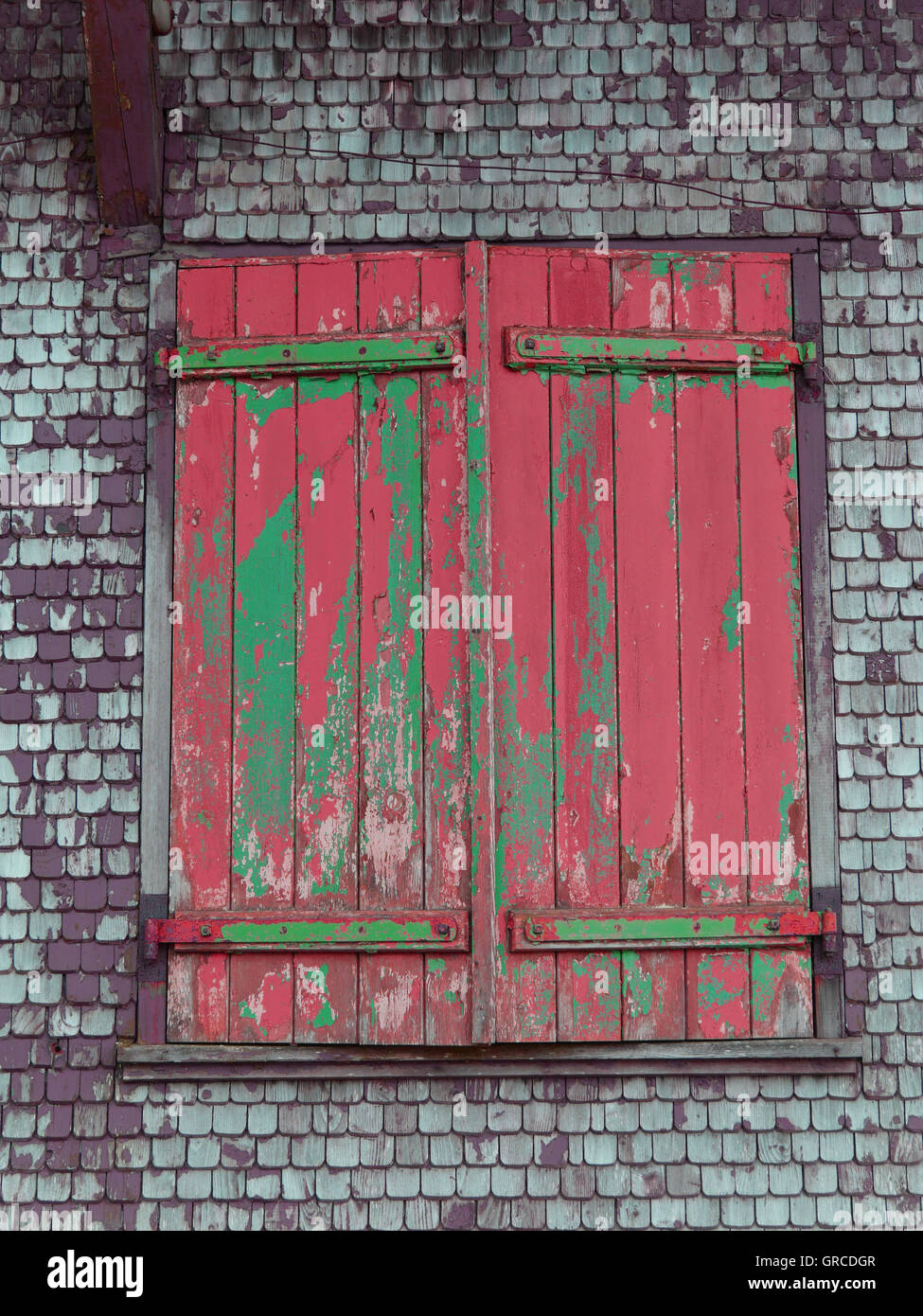 Closed Wooden Shutters With Exfoliated Red And Green Paint On Wall With Wooden Shingles Stock Photo