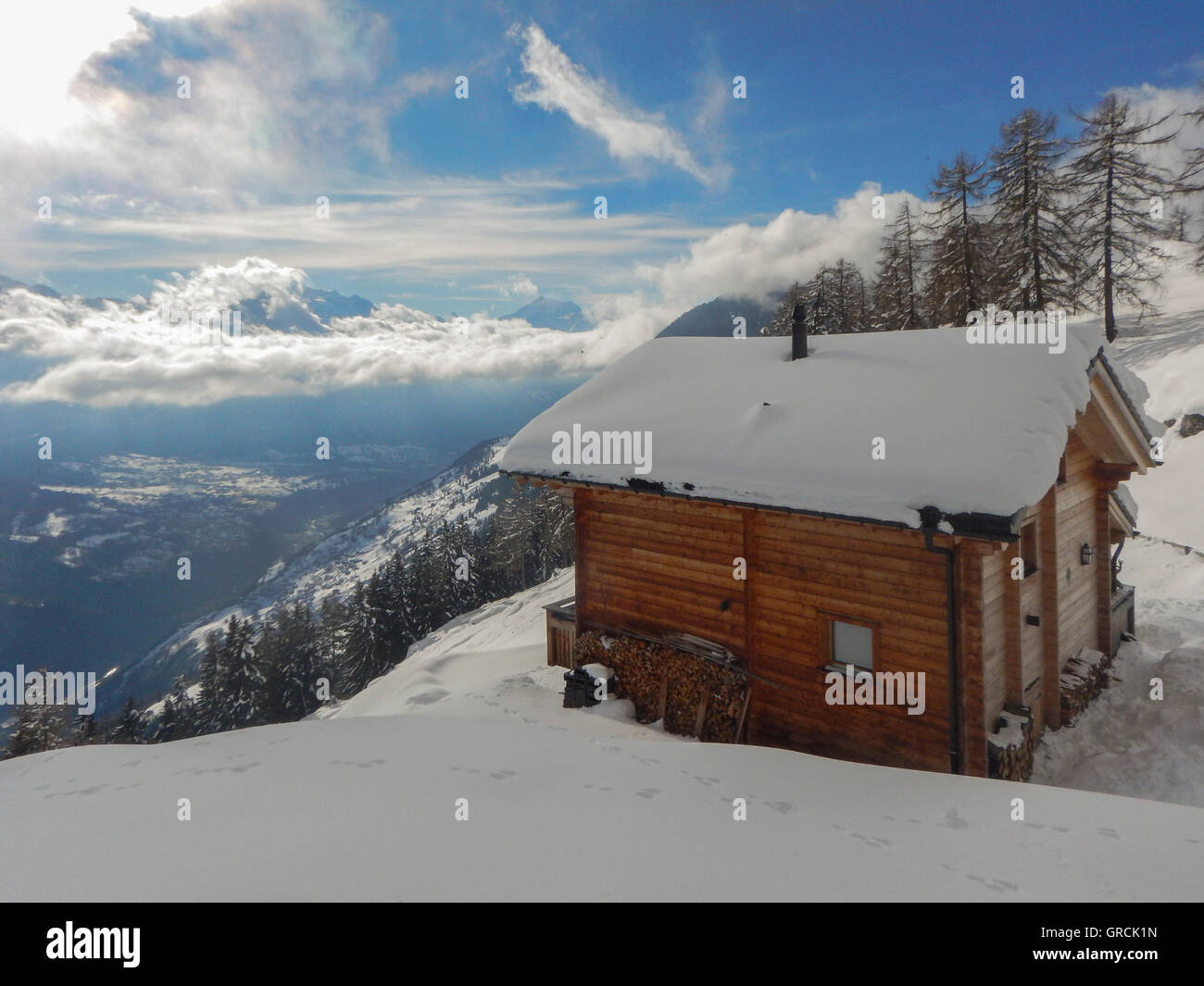 Snow Covered Chalet With A View To The Mountains Of The Valais As Wella Down Into The Valley. Blue Sky, Some Clouds. In The Foreground Snowcovered Slope. Sunny With Shadows. Stock Photo
