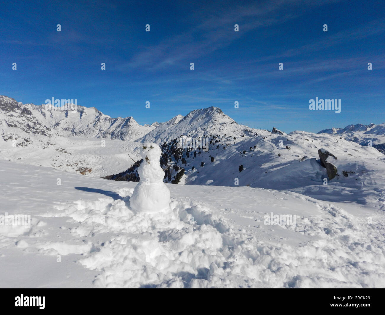 Snowman With Bettmerhorn And The Great Aletsch Glacier In The Background, On A Sunny Day In Winter With Deep Blue Sky And Snowcovered Mountains Stock Photo
