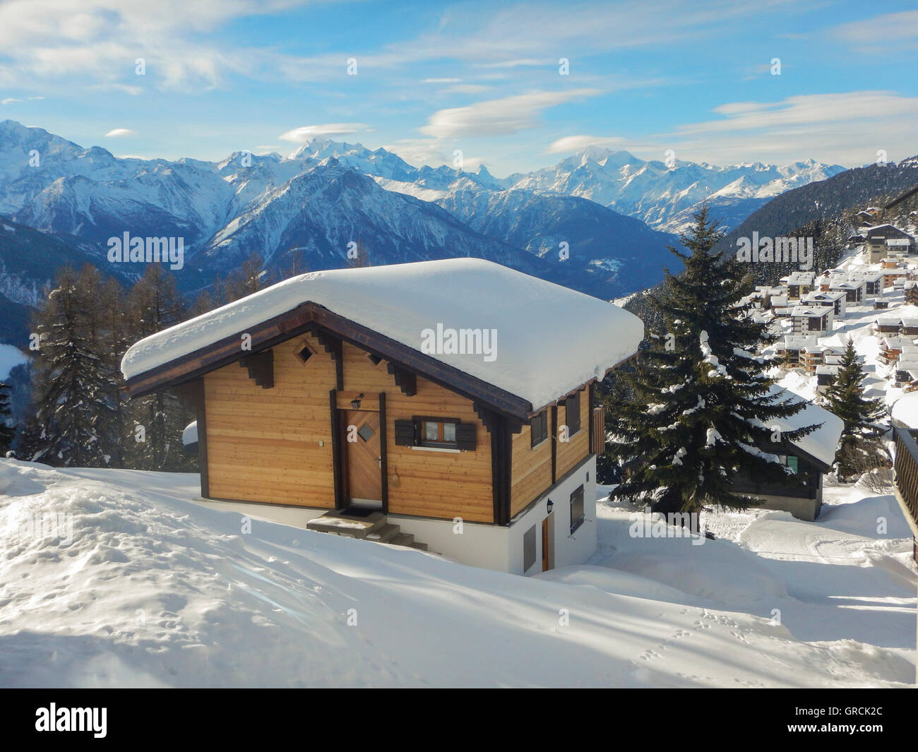 Snow Covered Chalet With A View To The Mountains Of The Valais. Blue Sky, Some Clouds. In The Foreground Snowcovered Hill. Sunny With Shadows. Stock Photo