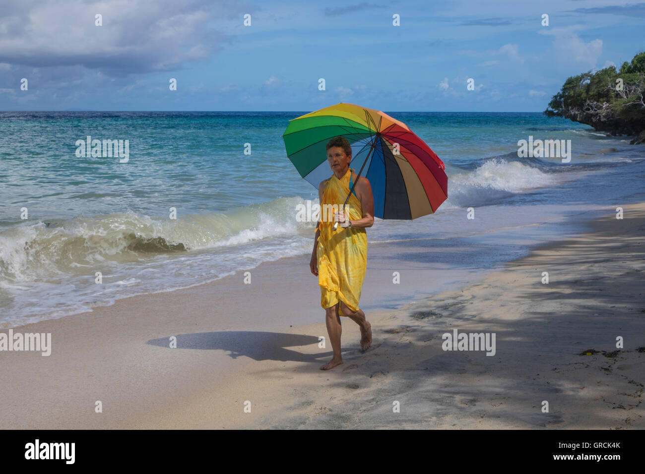 Woman In A Yellow Dress And A Rainbow Colored Parasol Walks Along A Sandy Beach. Stock Photo
