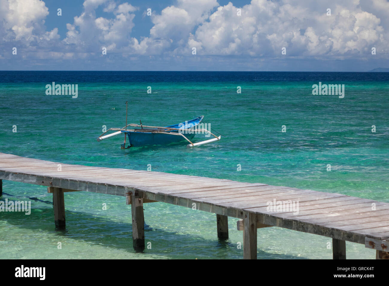 Indonesian Outrigger Canoe Near The Beach, Wooden Jetty In The Foreground, White Cumulus Clouds And Blue Sky In The Background Stock Photo
